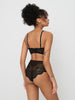 A woman modeling a black lace lingerie set from the back against a light gray studio background.