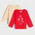Two long-sleeve children's shirts displayed on a light gray background. The front shirt is bright red with a Christmas-themed graphic print featuring a nutcracker, reindeer, candy cane, snowman, gingerbread man, and the text 'MAGIC TIME'.