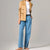 A model wearing light-wash blue straight-leg jeans, a white t-shirt, and a tan suede blazer, standing barefoot against a neutral gray studio background.
