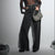 A model stands against a light gray studio background wearing a sheer black long-sleeve crop top over a black bandeau, high-waisted wide-leg dark gray jeans, and holding a silver sequined handbag.