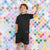 A young boy smiling and posing in a black athletic t-shirt and matching shorts set, standing against a colorful diamond-patterned tiled wall.
