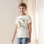 A young boy smiling and wearing a cream-colored short-sleeve t-shirt with a green and black dinosaur graphic on the front, paired with blue jeans, standing in front of a soft beige curtain background.