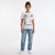 A woman modeling a white short-sleeve graphic t-shirt with a gold and black abstract design, paired with light-wash blue jeans and white sneakers, against a plain white studio background.