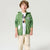 A young boy wearing a green zip-up hoodie over a white graphic t-shirt and beige pants, standing against a light gray studio background.