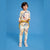 A young boy modeling a matching two-piece pajama set with a colorful abstract print on a cream base, featuring a long-sleeve top and pants, styled with a yellow tie belt and white sneakers against a solid blue background.