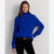 A woman wearing a bright royal blue cable-knit turtleneck sweater and black pants, standing against a light gray studio background.