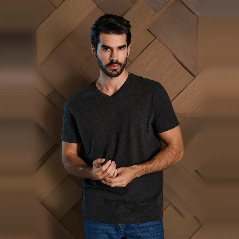 A man wearing a dark charcoal gray v-neck t-shirt and blue jeans, standing against a brown geometric textured background.