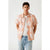 A man wearing a peach short-sleeve button-up shirt with a white palm leaf pattern, layered over a white t-shirt and light blue jeans, standing against a light gray studio background.