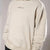 An oversized cream-colored hoodie with 'ALTERNATIVE' printed in black text on the chest, shown on a model against a neutral gray background.