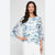 A woman modeling a light blue and white floral print blouse with three-quarter ruffled sleeves, paired with white pants, against a light gray studio background.