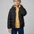 A young boy wearing a black hooded puffer jacket with a yellow lining, layered over a mustard yellow sweatshirt and blue jeans, photographed against a neutral gray studio background.