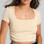 A woman wearing a beige short-sleeve crop top with a square neckline, paired with blue jeans and a black belt, shown from the chest down against a light gray background.
