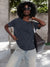 A woman wearing a dark charcoal gray t-shirt, light-wash blue jeans, sunglasses, and hoop earrings, carrying a dark brown leather shoulder bag in an outdoor urban setting.