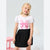 A young girl with blonde hair is wearing a white short-sleeve t-shirt with a pink bow graphic and a black pleated skirt, standing against a light gray background.