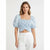 A woman modeling a light blue floral puff-sleeve crop top with a square neckline, paired with white pants, against a plain light gray studio background.