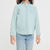 A young girl with long dark hair wearing a light blue chambray button-down shirt and light-wash jeans, standing against a plain light gray studio background.