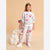 A young girl stands in a studio wearing a light gray two-piece pajama set with pink hearts and cat illustrations. She is posed next to a pink chair with a white blanket and a white flower-shaped pillow on a solid beige background.