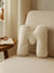 A fluffy cream-colored letter 'M' throw pillow resting on a textured beige armchair in a cozy, sunlit room.