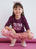 Young girl wearing a purple shirt with 'POSITIVE ENERGY' text and pink pants, sitting on a pink surface.