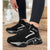 A person wearing black high-top sneakers with chunky silver and black geometric soles, standing on a textured beige floor. The shoes have black laces, a black ankle strap with a visible 'DAVID GEER' brand tag, and a black upper with silver graphic accents.