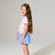 A young girl with long brown hair stands in profile against a light gray studio background, wearing a white and lavender striped short-sleeve top, a light blue skirt, and a small purple crossbody bag.