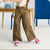 A child wearing leopard-print wide-leg pants, a white top, and red sneakers, standing in a bright indoor room with a desk and blue cabinet in the background.