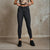 A model wearing dark gray slim-fit ankle-length jeans, a leopard-print crop top, and black leather shoes, standing against a textured concrete wall.
