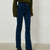 A person wearing dark blue high-rise straight-leg jeans, styled with an olive green knit sweater and black boots, standing against a plain light gray studio background.