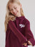 Young girl wearing a maroon hoodie with a logo on a plain background