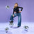 A young woman posing in a black long-sleeve top and wide-leg blue jeans, surrounded by four hanging and standing silver disco balls against a solid lavender-purple studio background.