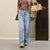 A person wearing light-wash blue straight-leg jeans, a brown top, an olive green blazer, and beige shoes, holding a beige handbag against a neutral indoor wall.
