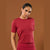A woman modeling a short-sleeve crewneck athletic top in a deep red color, paired with matching red bottoms, set against a solid warm brown studio background.