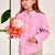 A young girl with long light brown hair wearing a pink button-up shirt and beige pants, holding a bouquet of orange and pink flowers next to a giant yellow flower prop against a plain light background.