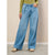 A model wearing light blue wide-leg denim jeans with a drawstring waist, paired with a green top and beige shoes, standing against a textured beige wall.