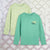 Two long-sleeve crewneck shirts displayed flat against a geometric white and pink background. The front shirt is mint green with a small 'APECK CROCODILE' graphic on the left chest, and the back shirt is pale yellow-green with a subtle reptile pattern.