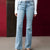 A person wearing light-wash blue flare jeans with a white long-sleeve top, shown from the waist down against a dark red and brown studio backdrop.