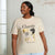 A woman wearing a beige graphic t-shirt with 'Classic Style' printed on the front, paired with light blue jeans, standing against a white brick wall.