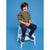 A young boy with brown hair sits smiling on a small white wooden step stool against a solid blue studio background. He is wearing an olive green long-sleeve henley shirt, dark navy pants, and navy sneakers with white laces.