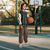 A young boy stands on an outdoor basketball court holding an orange basketball. He is wearing a dark green and beige varsity jacket, brown cargo pants, and green and white sneakers.