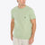 A man wearing a light sage green crewneck pocket t-shirt with a small black sailboat logo on the chest pocket, paired with off-white shorts, against a plain white background.