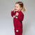 A young girl with blonde hair in a ponytail smiles while wearing a matching burgundy red two-piece pajama set with cute animal appliques, photographed against a neutral gray studio background.