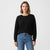A woman wearing a black long-sleeve crewneck sweater and blue jeans, standing against a plain white studio background.