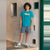 A child model wearing a bright turquoise graphic t-shirt with 'VIBES' and a hand graphic, paired with slate blue shorts, white socks, and green and white sneakers, standing in a doorway with textured white walls.