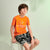 A boy sitting on a white stool wearing a bright orange Las Palmas t-shirt, black and white patterned shorts, and black sneakers against a light green studio background.