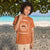 A child wearing a terracotta orange graphic t-shirt with a white sun design and 'FLORIDA' text, standing on a sandy beach with turquoise water and a tree canopy in the background.