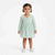 A toddler girl wearing a mint green long-sleeve tulle dress with a crossover V-neck and a small bow detail at the waist, standing against a plain white studio background.