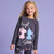 A young girl with curly blonde hair smiling while wearing a dark gray long-sleeve pajama top and matching patterned pajama pants featuring Stitch and Angel graphics, set against a solid lavender studio background.