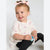 A toddler girl sitting on a light gray studio floor, wearing a cream long-sleeve top with a pink bow and butterfly print, black leggings, and a pink bow in her hair, holding a small blue toy.