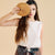 A woman modeling a sleeveless white top and brown skirt, holding a large round wooden slice in front of her face. She is wearing a dark belt with silver studs and a decorative hanging strap against a solid beige background.