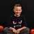 A smiling young boy sits cross-legged on a black fabric backdrop, wearing a black long-sleeve T-shirt with a cartoon monster face graphic featuring red eyes and white fangs. Halloween props, including a pumpkin, a jack-o'-lantern bucket, and popcorn, are visible around him.